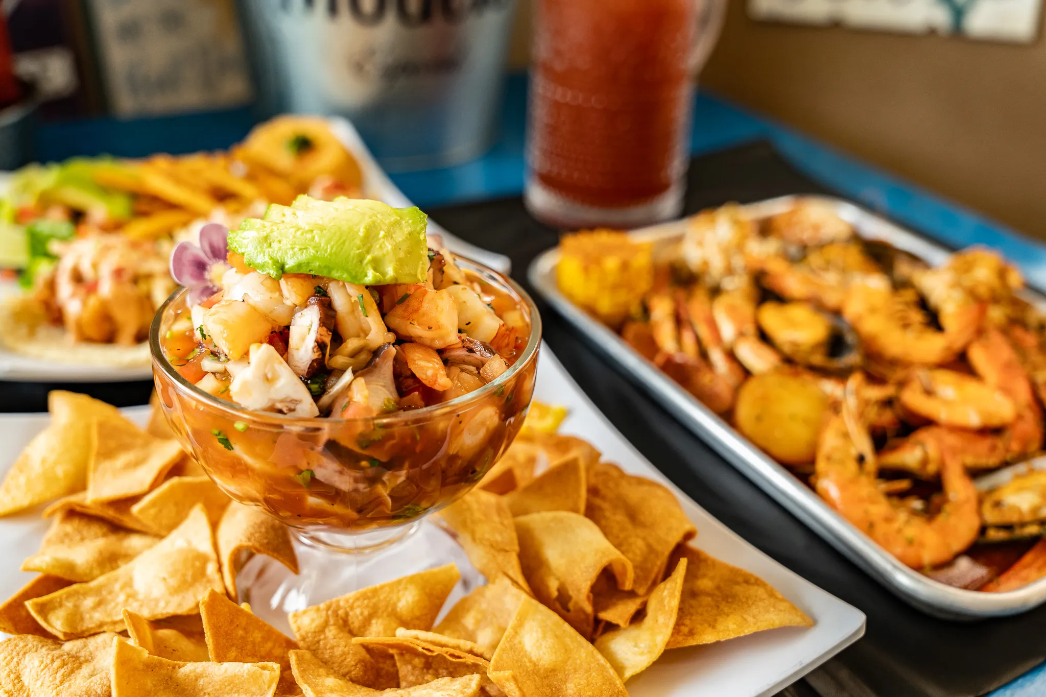 Shrimp ceviche in a footed glass with a bloody mary and a plate of grilled seafood in the background