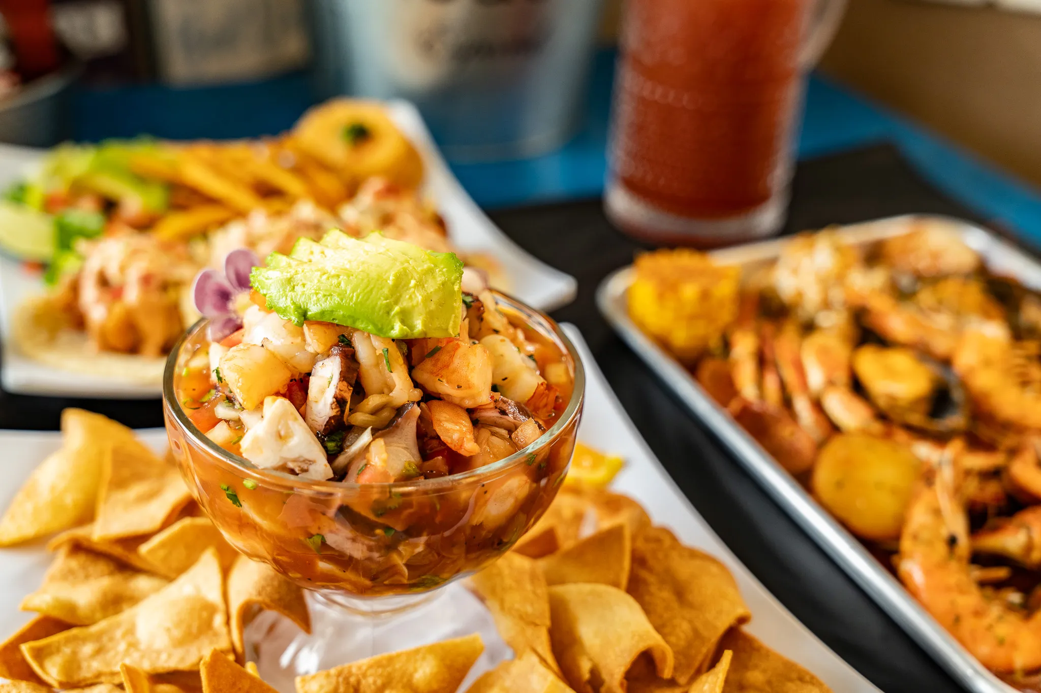 A glass bowl of shrimp ceviche topped with avocado on a bed of tortilla chips, beside a tray of seafood boil at Wacky Crab Shack