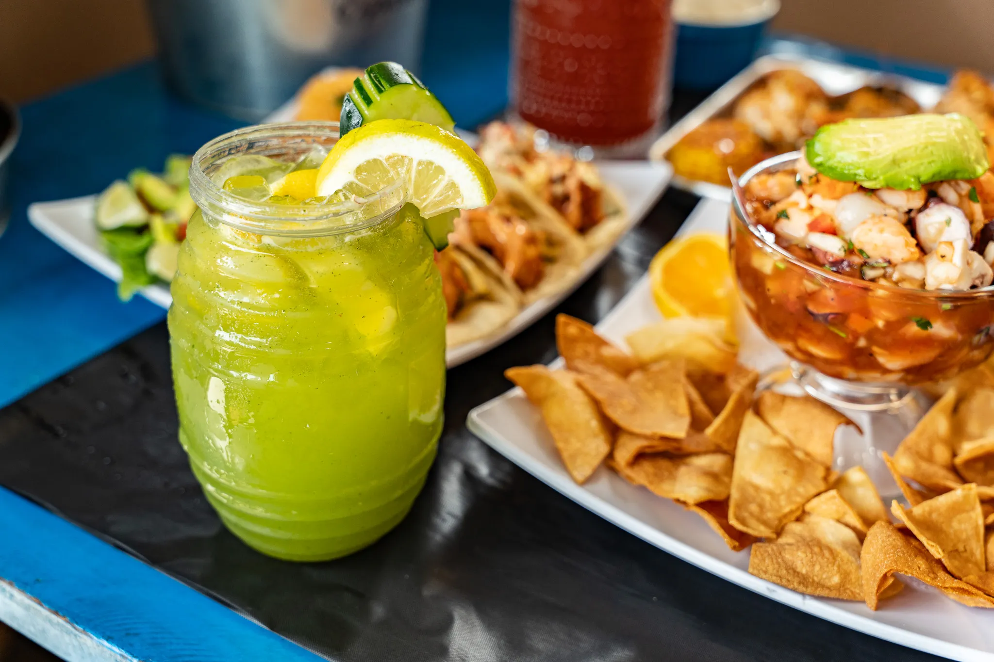 A bright green Wacky Crab Shack cocktail beside a plate of shrimp tacos and a bowl of ceviche